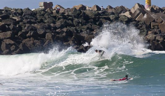 Surfer riding a wave beside the rocky breakwater at La Barre